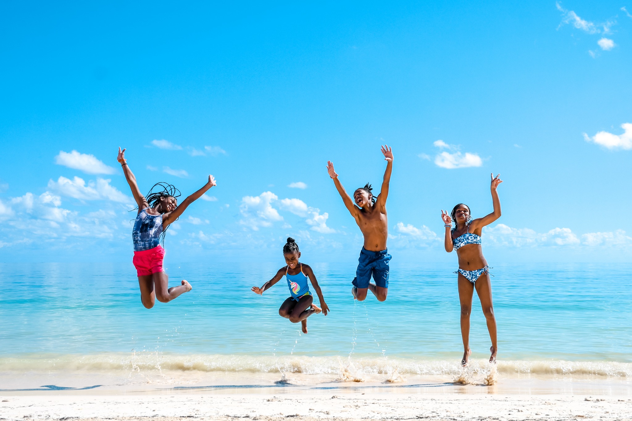 Four children jumping on a sunny beach beside turquoise water.